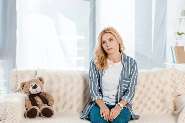 Upset woman sitting on sofa near soft toy at home — Stock Photo