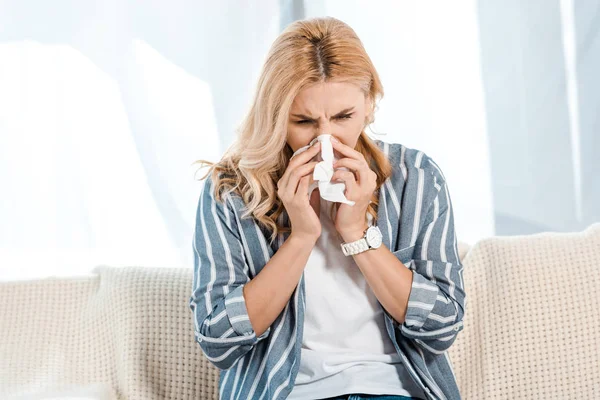 Kranke Frau sitzt auf Sofa und niest in Serviette — Stockfoto