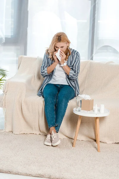 Больная женщина сидит на диване и чихает в салфетке рядом с коробкой ткани — стоковое фото