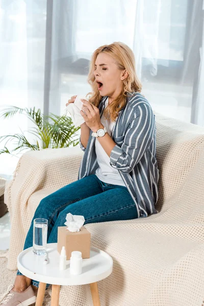 Frau mit geschlossenen Augen sitzt auf Sofa und niest, während sie Serviette hält — Stockfoto
