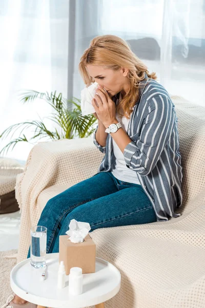 Woman with closed eyes sitting on sofa and sneezing while holding tissue — Stock Photo