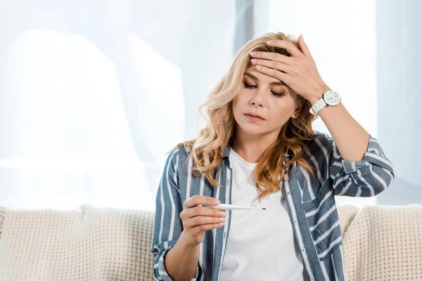 Kranke Frau berührt Stirn und blickt auf elektronisches Thermometer — Stockfoto