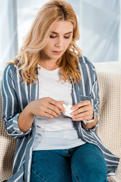 Attractive woman looking at bottle with pills while sitting on sofa — Stock Photo