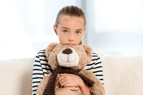 Niedliches Kind hält Teddybär in der Hand, während es im Wohnzimmer in die Kamera schaut — Stockfoto