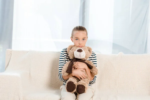 Niedliches Kind hält Teddybär, während es in die Kamera schaut und auf dem Sofa sitzt — Stockfoto