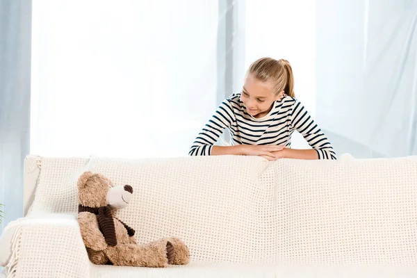 Niño feliz mirando osito de peluche en el sofá - foto de stock