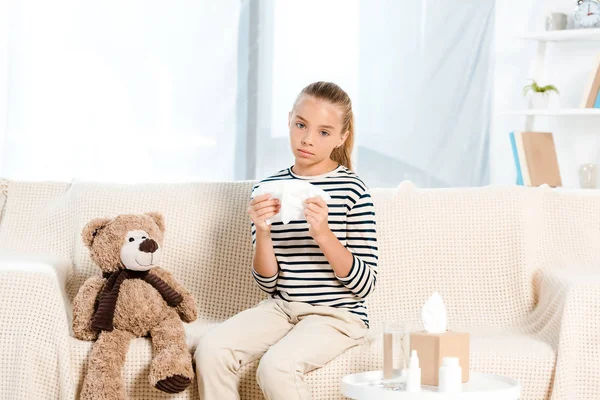 Sick kid holding napkin near teddy bear and sitting on sofa — Stock Photo