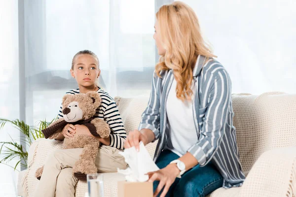 Kid with teddy bear looking at sick mother taking tissue at home — Stock Photo
