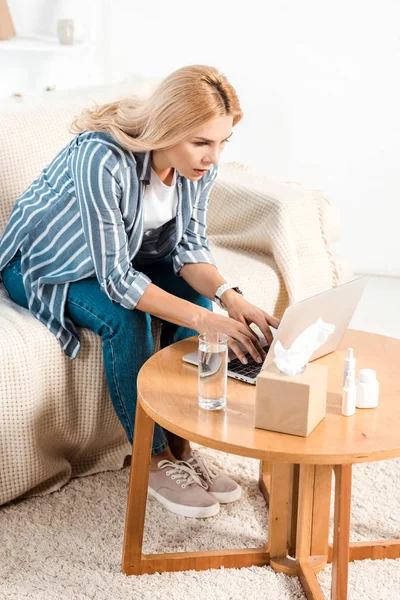 Atractiva mujer usando portátil cerca de vidrio de agua y caja de tejido en casa - foto de stock