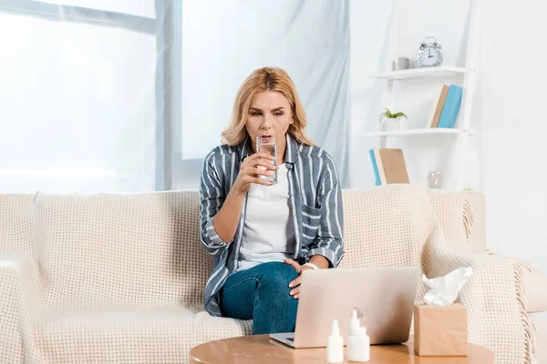 Sick woman drinking water while looking at laptop near tissue box at home — Stock Photo