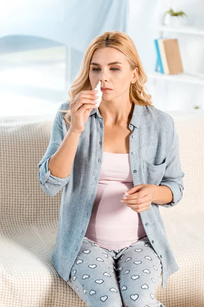 Mulher grávida doente usando spray nasal enquanto sentada no sofá — Fotografia de Stock