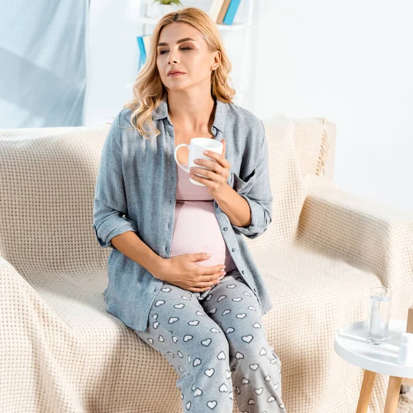 Attractive and pregnant woman sitting on sofa with cup of tea — Stock Photo