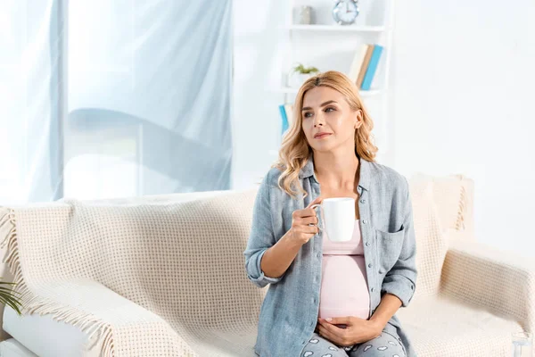 Schöne schwangere Frau sitzt auf dem Sofa und hält Tasse mit Tee — Stockfoto