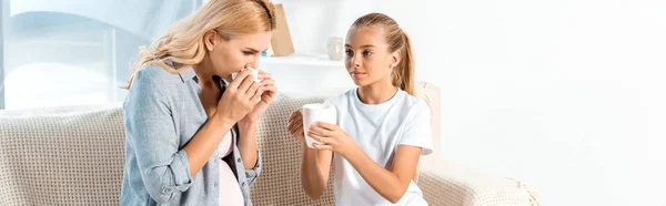 Panoramic shot of kid holding cup near mother sneezing in tissue — Stock Photo