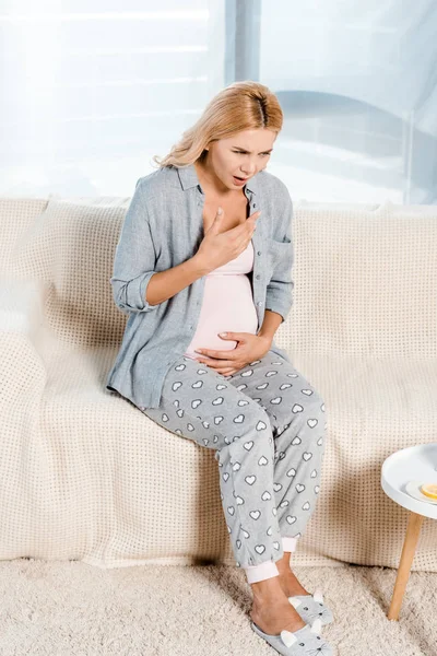 Mujer embarazada enferma sentada en el sofá en la sala de estar - foto de stock