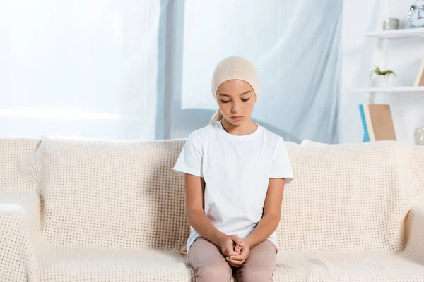 Upset kid in head scarf sitting on sofa in living room — Stock Photo