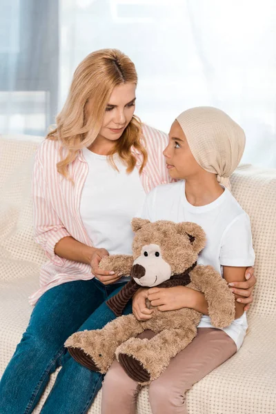 Atractiva madre sentada en el sofá con el niño enfermo con oso de peluche - foto de stock