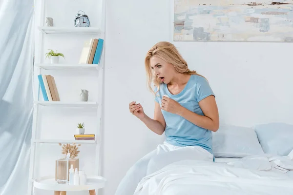 Mulher chocada segurando termômetro digital enquanto sentado na cama em casa — Fotografia de Stock