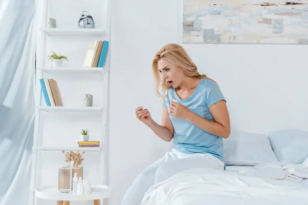 Shocked woman holding digital thermometer while sitting on bed at home — Stock Photo