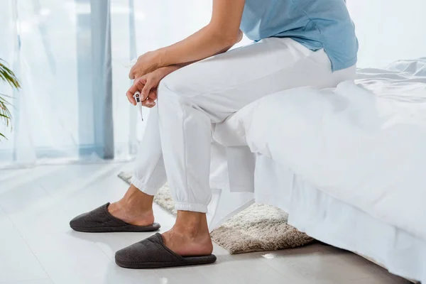 Cropped view of woman holding digital thermometer while sitting on bed — Stock Photo