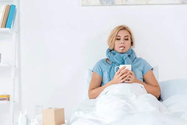 Mujer enferma en pañuelo azul sosteniendo termómetro digital en la boca y taza en las manos - foto de stock