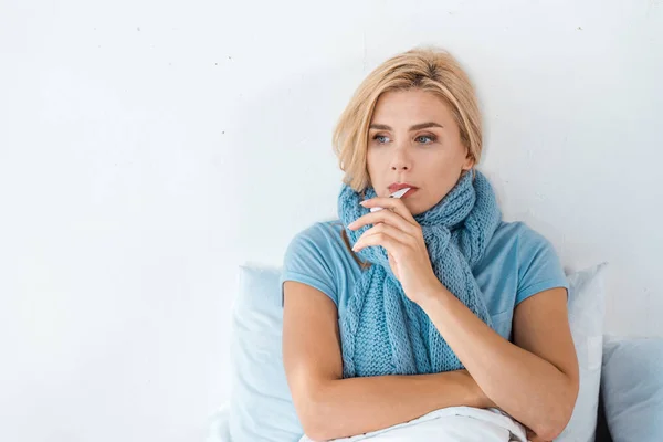 Kranke Frau mit blauem Schal hält digitales Thermometer im Mund im Schlafzimmer — Stockfoto