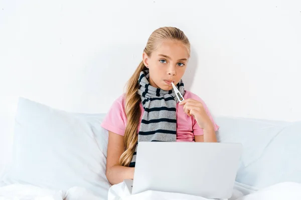 Niño enfermo sosteniendo termómetro digital en la boca cerca del ordenador portátil - foto de stock