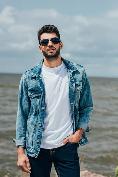 Hombre guapo en chaqueta de mezclilla y gafas de sol mirando a la cámara - foto de stock