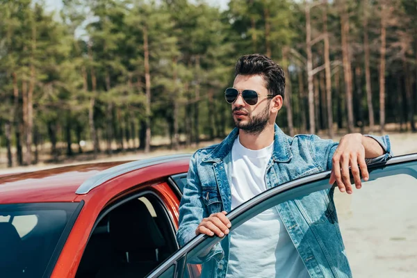 Красивий чоловік в сонцезахисних окулярах і джинсовій куртці, що стоїть біля машини — стокове фото