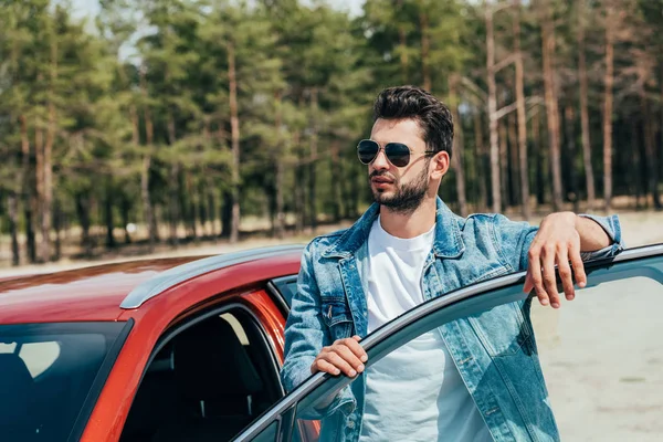 Hombre guapo en gafas de sol y chaqueta de mezclilla de pie cerca del coche - foto de stock