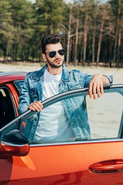 Bel homme en lunettes de soleil et veste en denim debout près de la voiture — Photo de stock