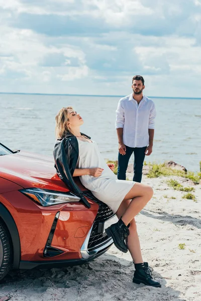 Attractive woman in dress with closed eyes lying on car and man look at her — Stock Photo