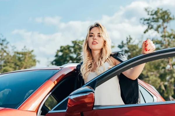 Attraktive und blonde Frau, die neben Auto steht und in die Kamera schaut — Stockfoto