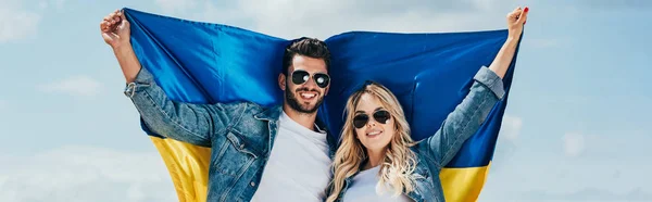 Panoramic shot of attractive woman and handsome man smiling and holding ukrainian flag — Stock Photo