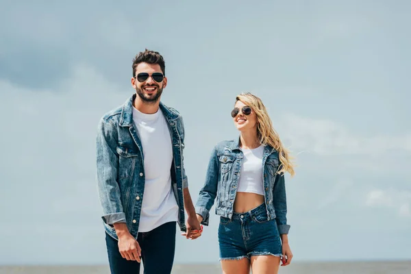 Mujer atractiva y hombre guapo en chaquetas de mezclilla sonriendo y tomándose de las manos fuera - foto de stock