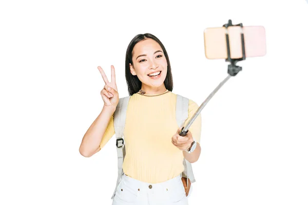 Attractive asian woman showing victory sign while taking selfie on smartphone with selfie stick isolated on white — Stock Photo