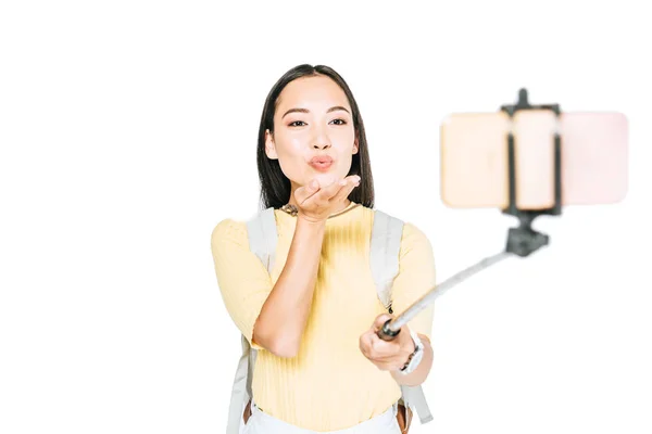 Attractive asian woman sending air kiss while taking selfie on smartphone with selfie stick isolated on white — Stock Photo