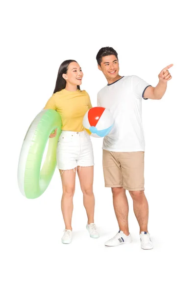 Bel homme asiatique avec boule de plage pointant du doigt tout en se tenant près de la petite amie avec anneau de natation sur fond blanc — Photo de stock