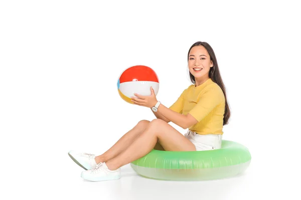 Cheerful asian girl sitting on swim ring and holding beach ball on white background — Stock Photo