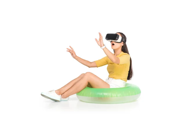 Schockierte asiatische Frau mit Virtual-Reality-Headset und gestikuliert, während sie auf Schwimmring auf weißem Hintergrund sitzt — Stockfoto