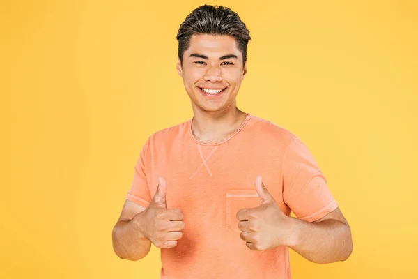 Guapo asiático hombre mostrando pulgares arriba y sonriendo a cámara aislado en amarillo - foto de stock