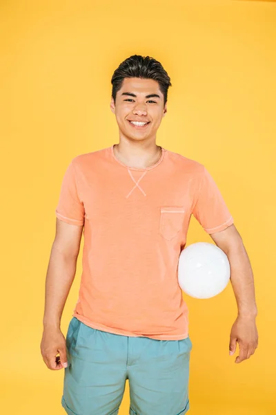 Jovem asiático homem segurando bola e sorrindo para câmera isolado no amarelo — Fotografia de Stock