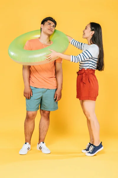 Happy asian girl putting swimming ring on smiling boyfriend on yellow background — стоковое фото