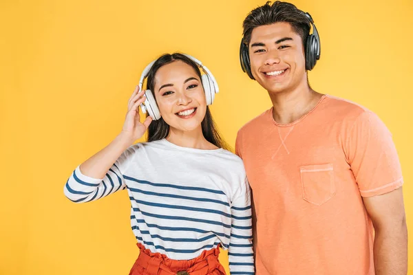 Cheerful asian man and woman listening music in headphones while smiling at camera isolated on yellow — Stock Photo