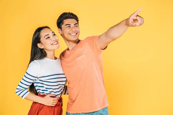 Happy asian man looking away and pointing with finger while standing near smiling girlfriend isolated on yellow — Stock Photo