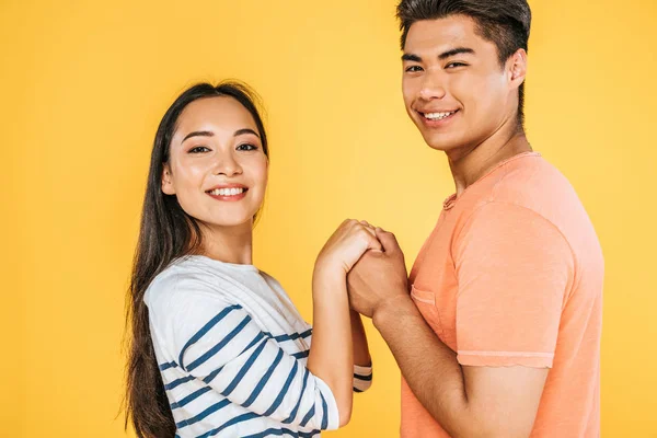 Felice asiatico uomo e donna tenendosi per mano mentre guardando fotocamera isolato su giallo — Foto stock