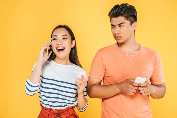 Cheerful asian girl talking on smartphone near dissatisfied boyfriend isolated on yellow — Stock Photo