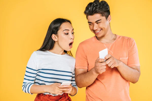 Smiling asian man showing smartphone to shocked girlfriend isolated on yellow — Stock Photo