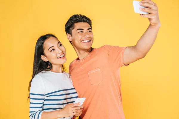Feliz asiático hombre tomando selfie en smartphone con hermosa novia aislado en amarillo - foto de stock