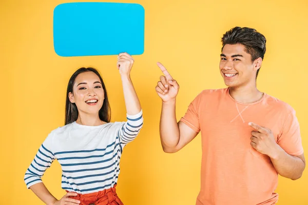 Asian man pointing with finger at speech bubble in hands of smiling girlfriend on yellow background — Stock Photo